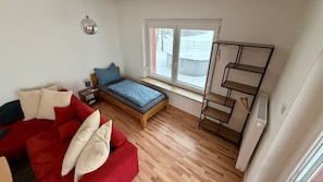 Una habitación compacta con una cama, un sofá rojo, una mesita, un aplique colgante, un ventanal con vistas y un radiador.