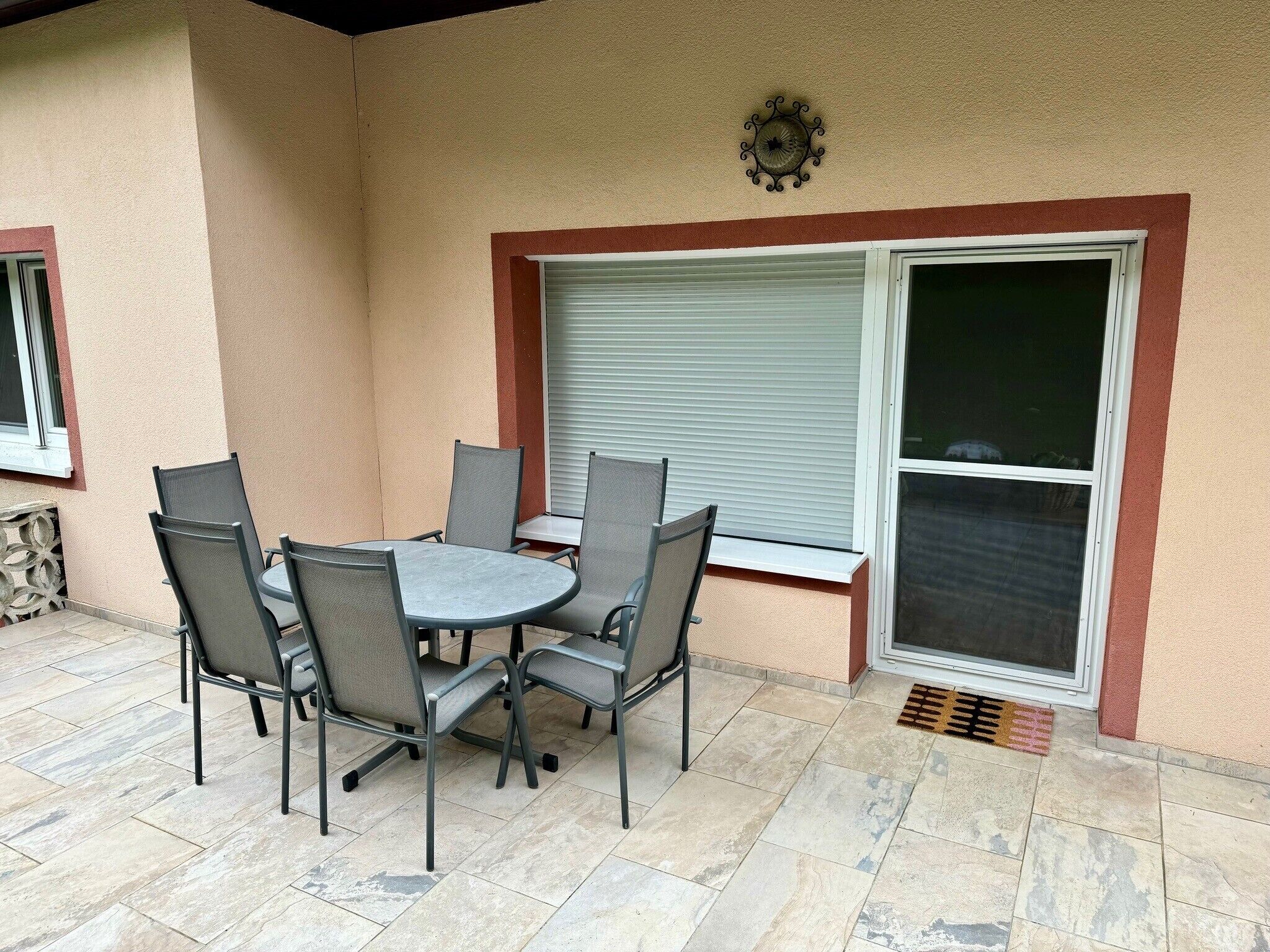Un patio al aire libre con una mesa redonda y seis sillas, una ventana con persianas y una puerta con mosquitero.