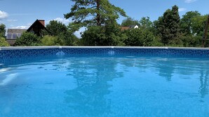 Une piscine d’un bleu éclatant, bordée de carreaux, entourée d’arbres et avec une maison en arrière-plan.