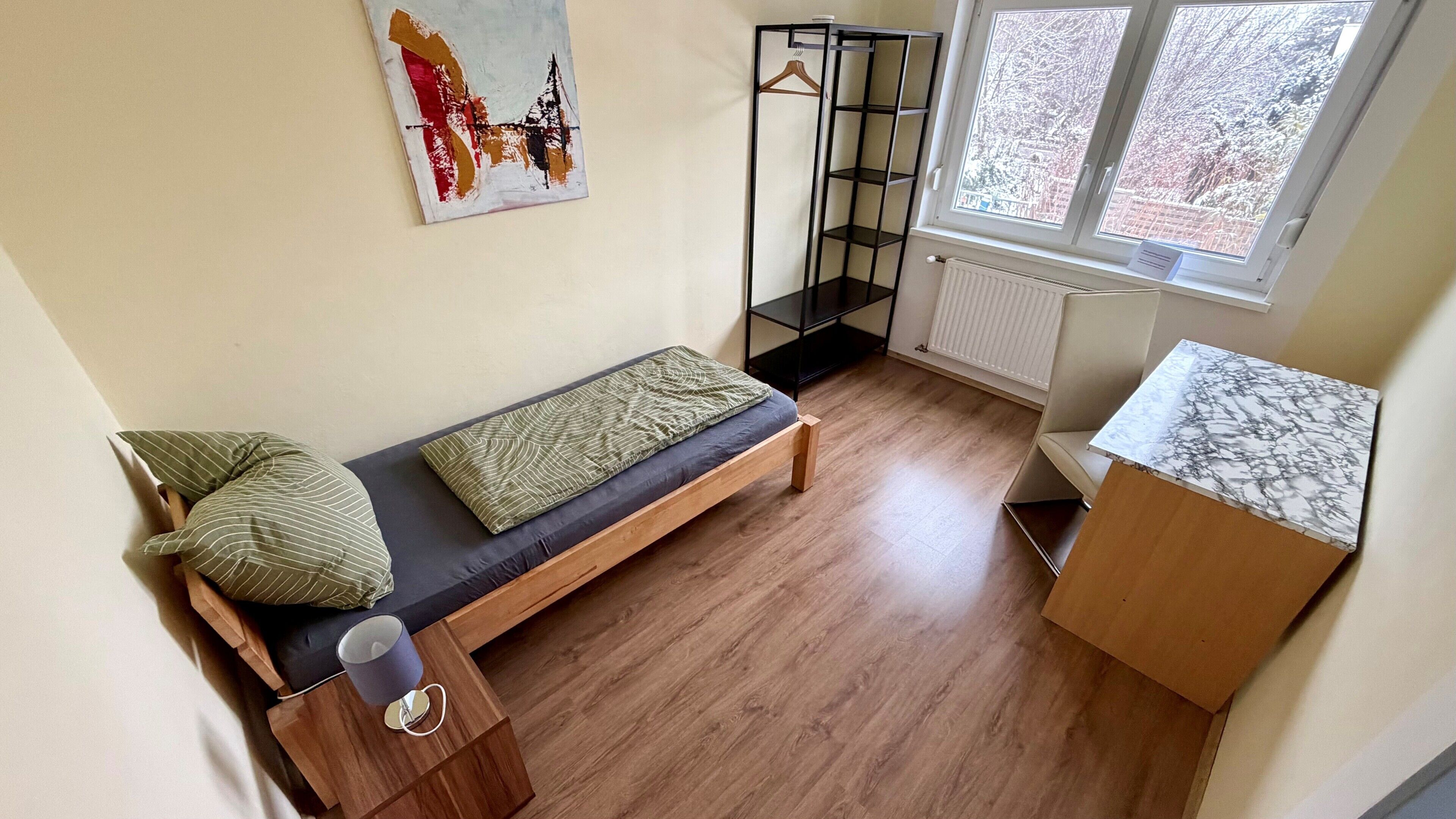 Een kleine, praktische kamer met een bed, een nachtkastje met lamp, een radiator, een raam en een plank.