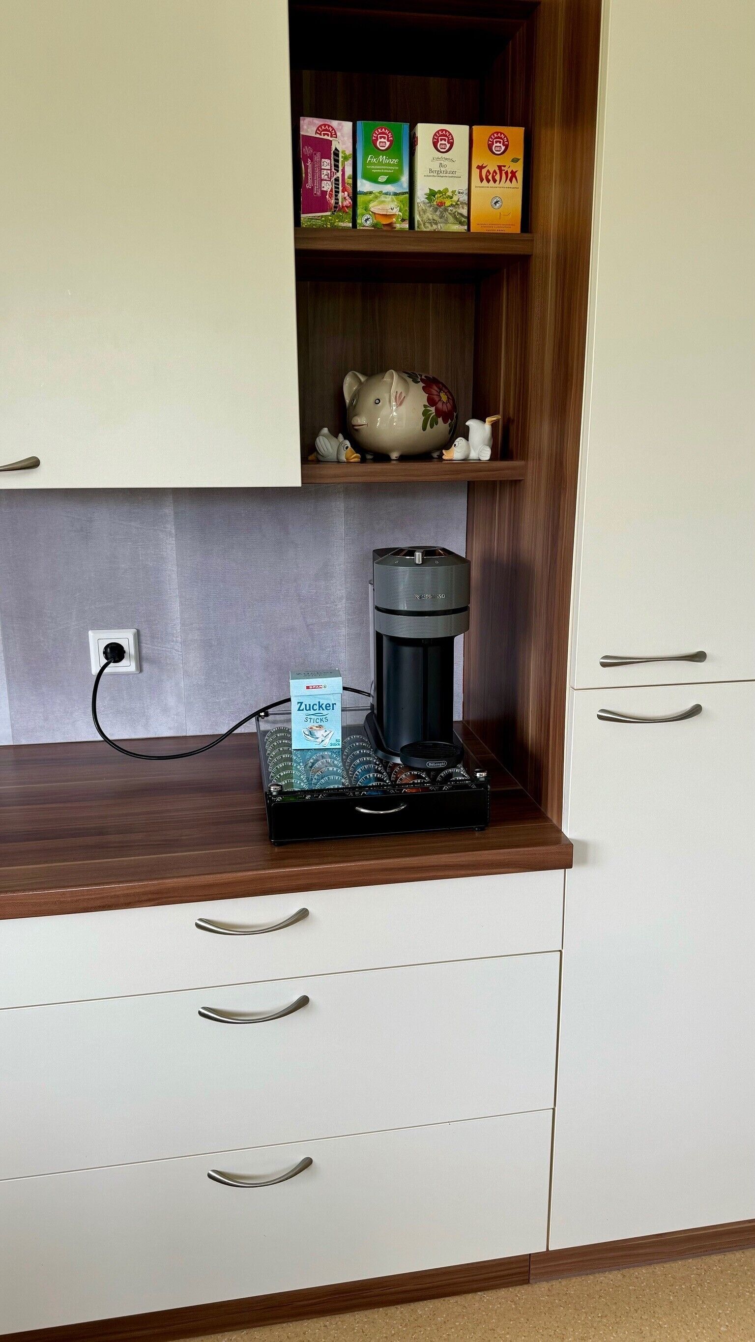Ett litet kök med en kaffebryggare, en förpackning kaffekapslar och en spargris på hyllan.