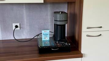 Ett litet kök med en kaffebryggare, en förpackning kaffekapslar och en spargris på hyllan.