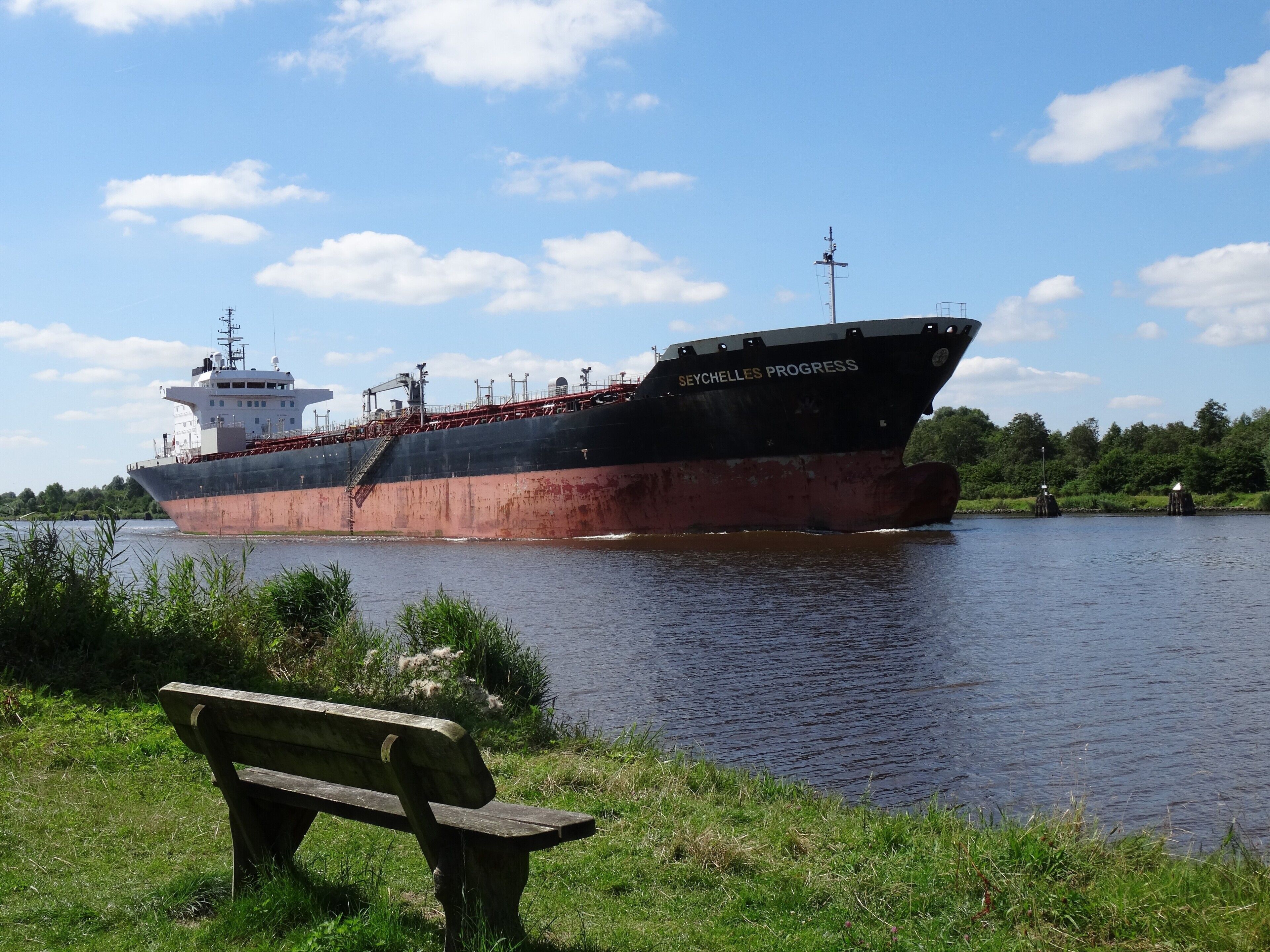 A large cargo ship named 'SEYCHELLES PROGRESS' docked at a port with a grassy area and a wooden bench in the foreground.
