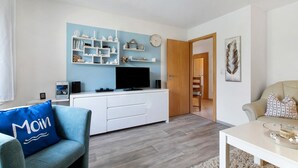 A modern living room with a white entertainment unit, a flat-screen TV, and a blue sofa with a 'Moin' pillow. - Holiday Apartment Auszeit an der Eider – Relax in Idyllic Hennstedt (Hennstedt)