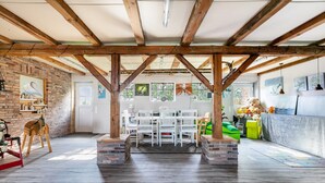 A spacious room with exposed wooden beams, a brick wall, and a dining area with white chairs and a table. - Apartment 'Ferienwohnung Hof Krakebüll' with Private Terrace, Private Garden and Wi-Fi (Humptrup)