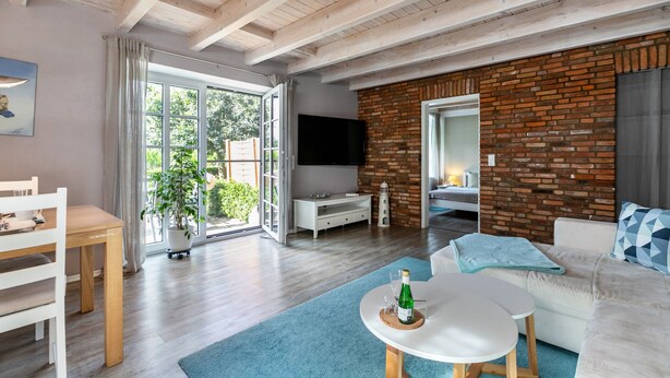 A modern living room with a wooden ceiling, a brick wall, a flat-screen TV, a white sofa, a dining table, and a large window. - Apartment 'Ferienwohnung Hof Krakebüll' with Private Terrace, Private Garden and Wi-Fi (Humptrup)