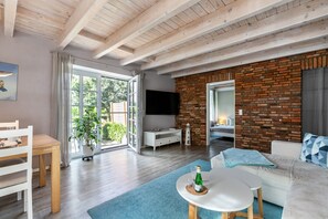 A modern living room with a wooden ceiling, a brick wall, a flat-screen TV, a white sofa, a dining table, and a large window. - Apartment 'Ferienwohnung Hof Krakebüll' with Private Terrace, Private Garden and Wi-Fi (Humptrup)