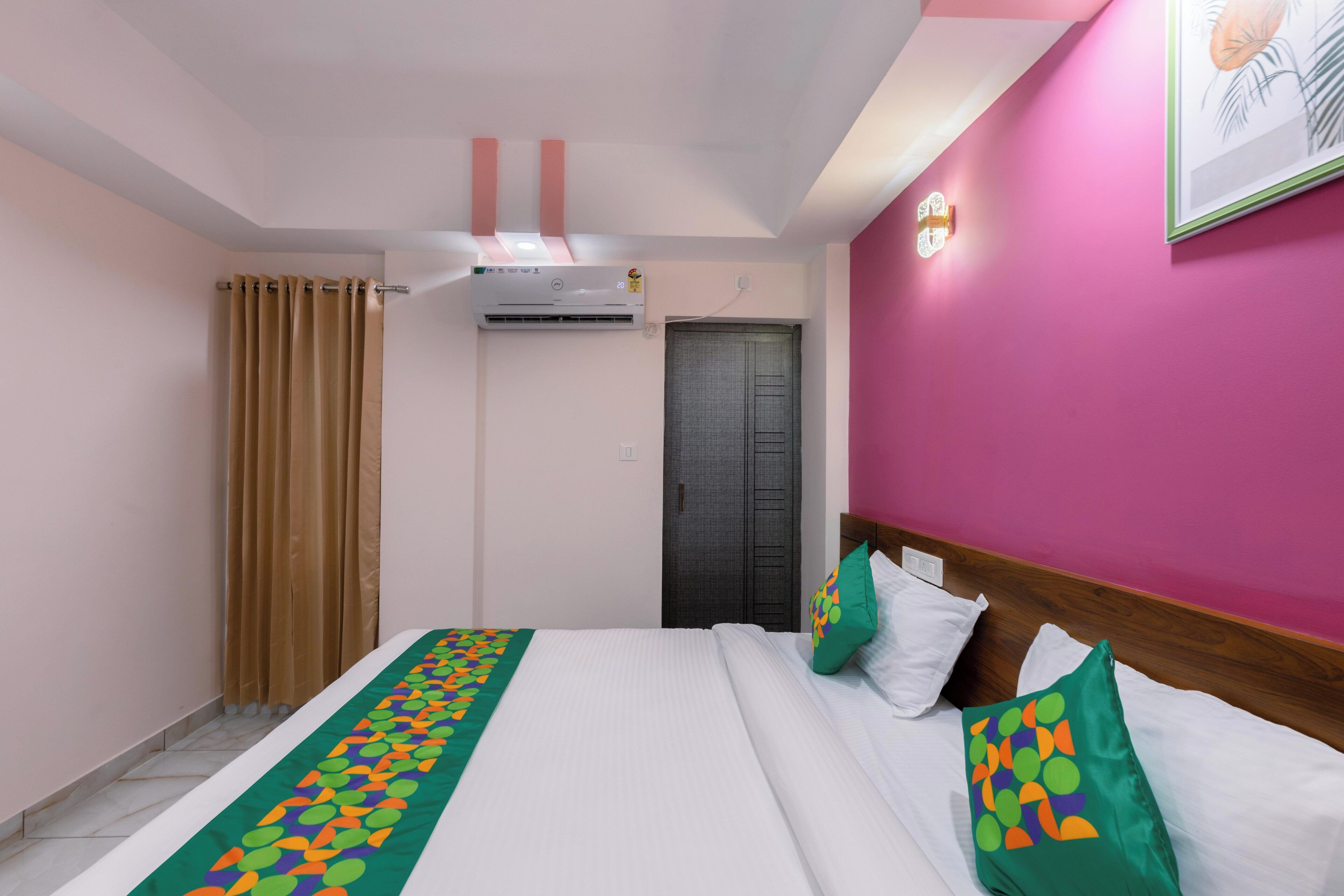 A hotel room with a bed, a curtain, an air conditioning unit, a door, and a wall with a light switch.