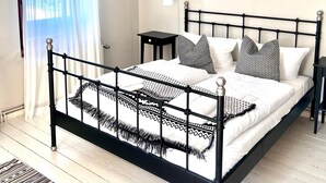 Een slaapkamer met een zwart metalen bed, wit beddengoed, een klein nachtkastje en een plafondlamp.