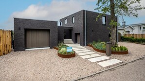 A modern two-story building with a dark brick exterior, a garage, and a gravel driveway leading to a wooden fence. - Attractive villa with jetty (Kamperland)