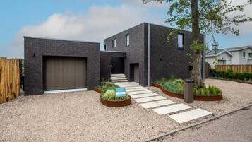 Un bâtiment moderne à deux étages, doté d’une façade en briques sombres, d’un garage et d’une allée en gravier menant à une clôture en bois.