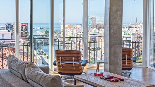 A modern living room with a sofa and two wooden chairs, a coffee table, and a view of a cityscape through large windows. - Solaga - Linaje (Málaga)