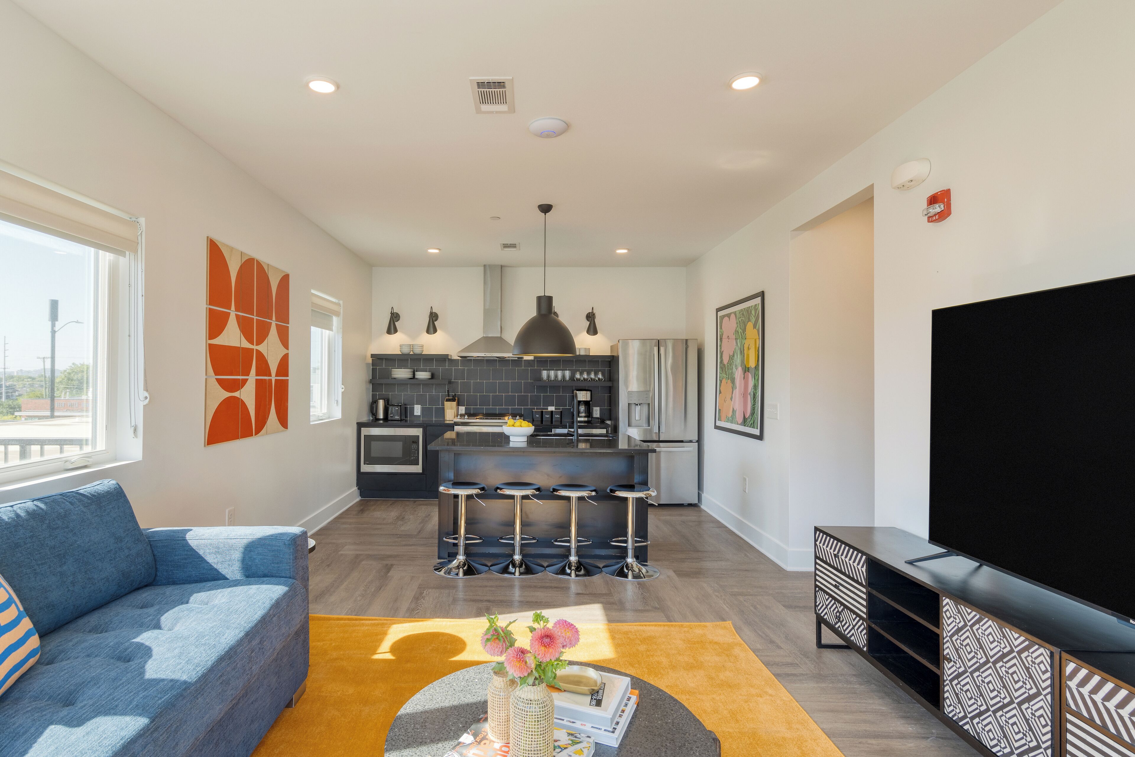 A modern living room with a sofa, a coffee table with decorative items, a kitchen area with a central island and bar stools, and a large flat-screen TV.