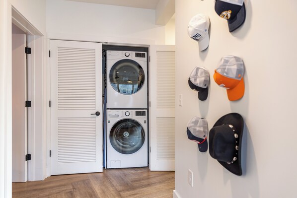 A laundry room with two stacked washing machines and a wall-mounted hat rack with various hats. - Bachelor Party Pad, 5 Minutes to Broadway + Private Balcony | Last Call (Nashville)