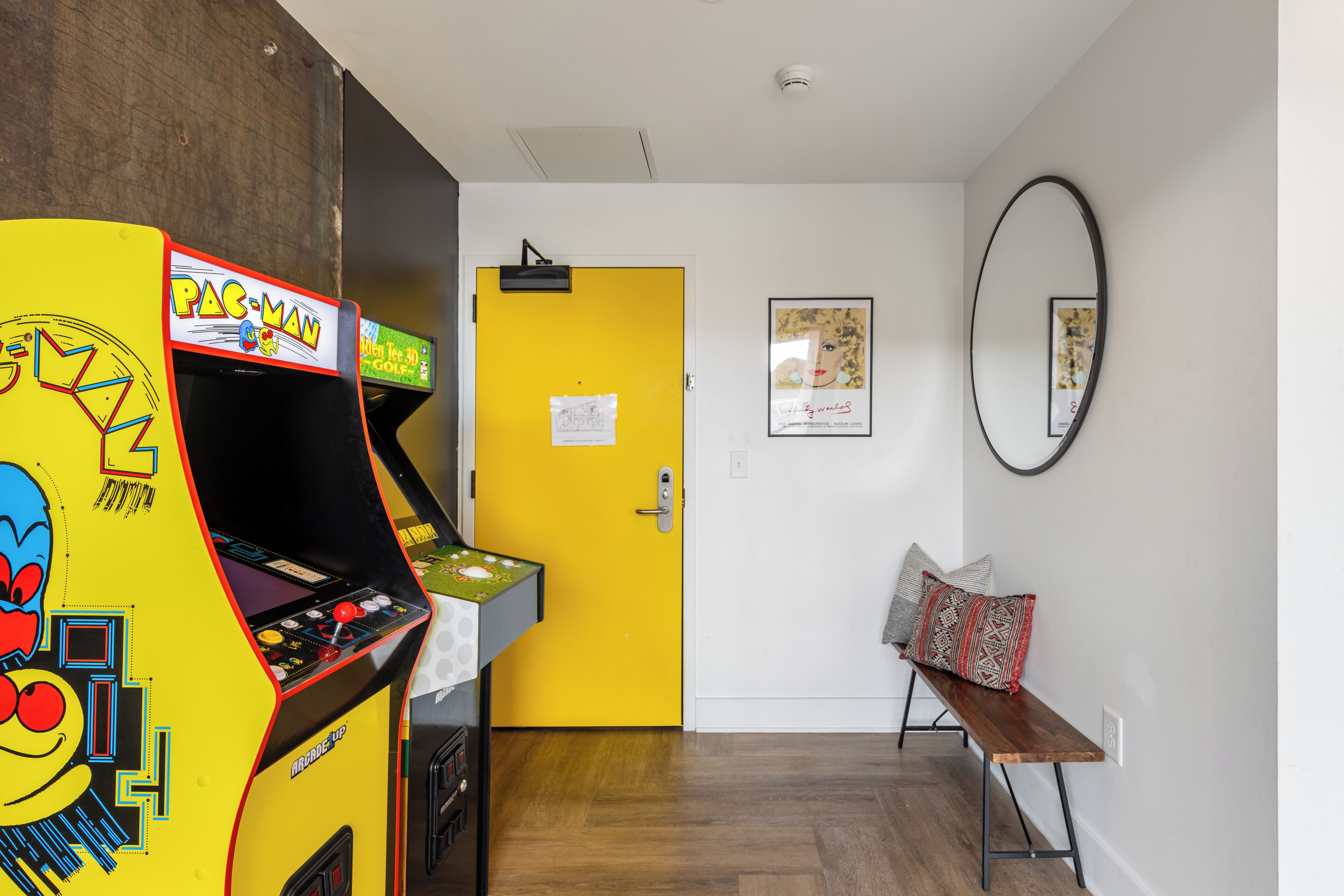 A room with a Pac-Man arcade machine, a yellow door, a bench with pillows, and a wall-mounted mirror.