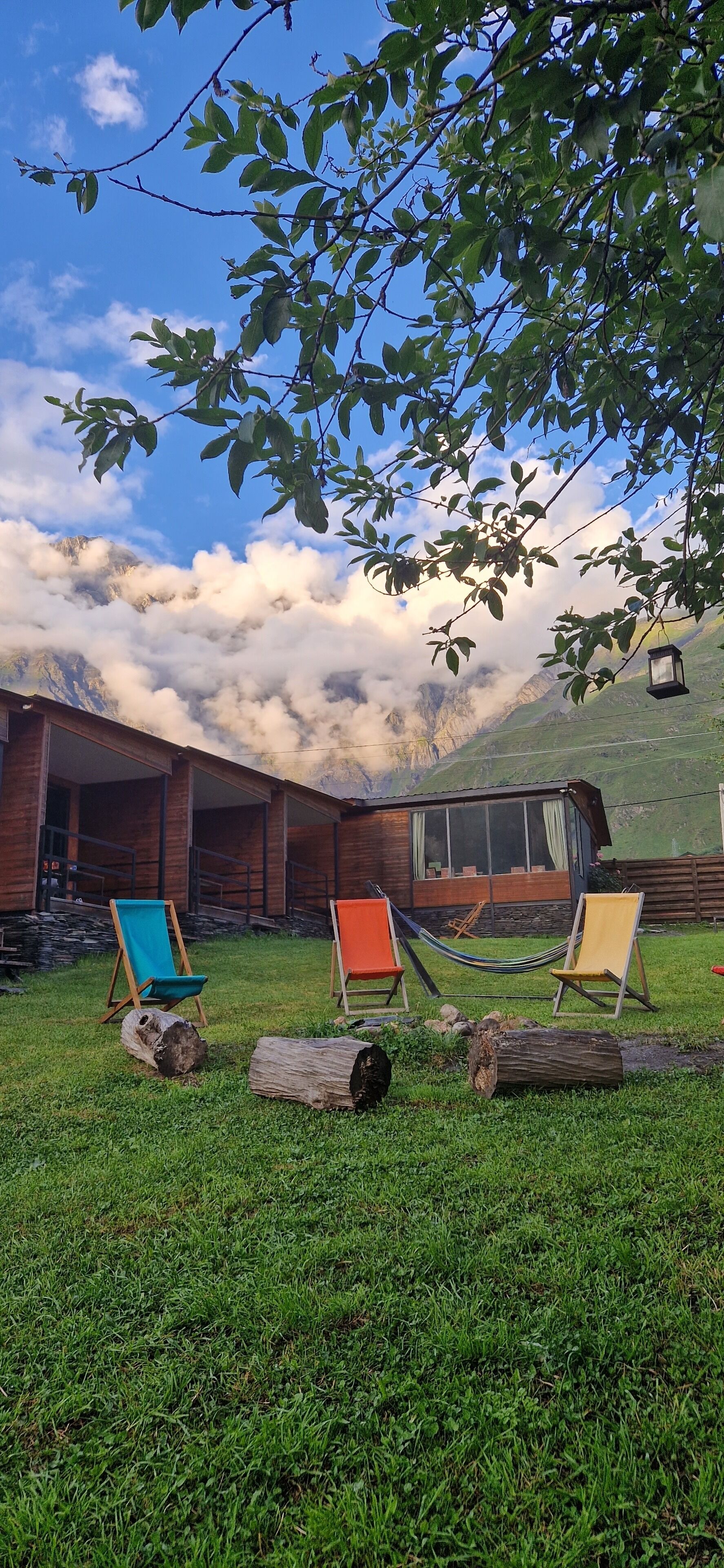 Una cabaña de madera con puerta de vidrio, tres sillas coloridas, una hamaca y un banco de troncos en un área cubierta de césped, con montañas al fondo.