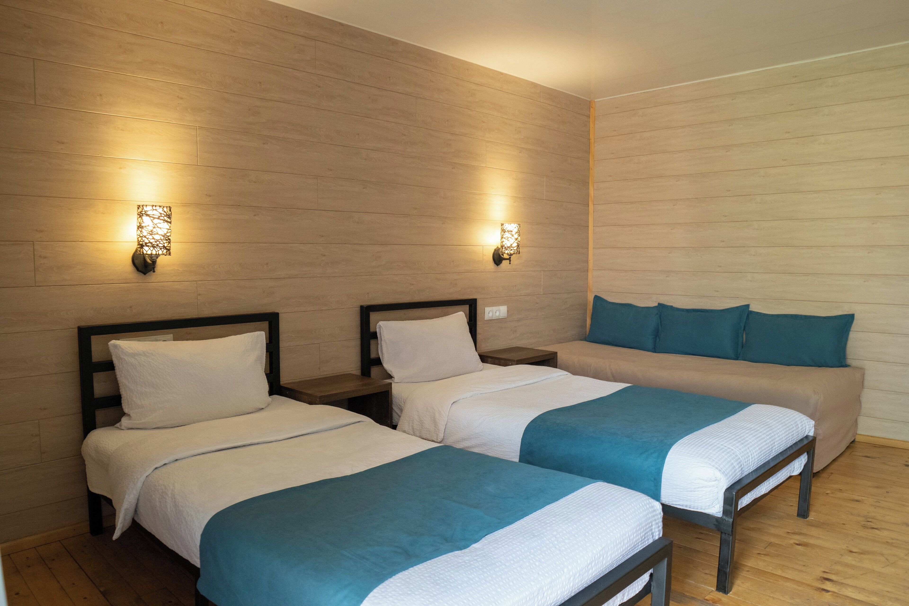 A hotel room with two single beds and a sofa bed, wooden panel walls, and wall-mounted lighting.