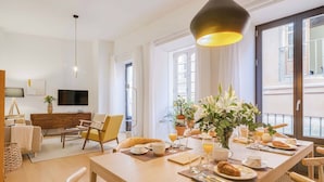 Un'area pranzo moderna con un tavolo in legno apparecchiato per la colazione, una TV sullo sfondo e un balcone con vista.