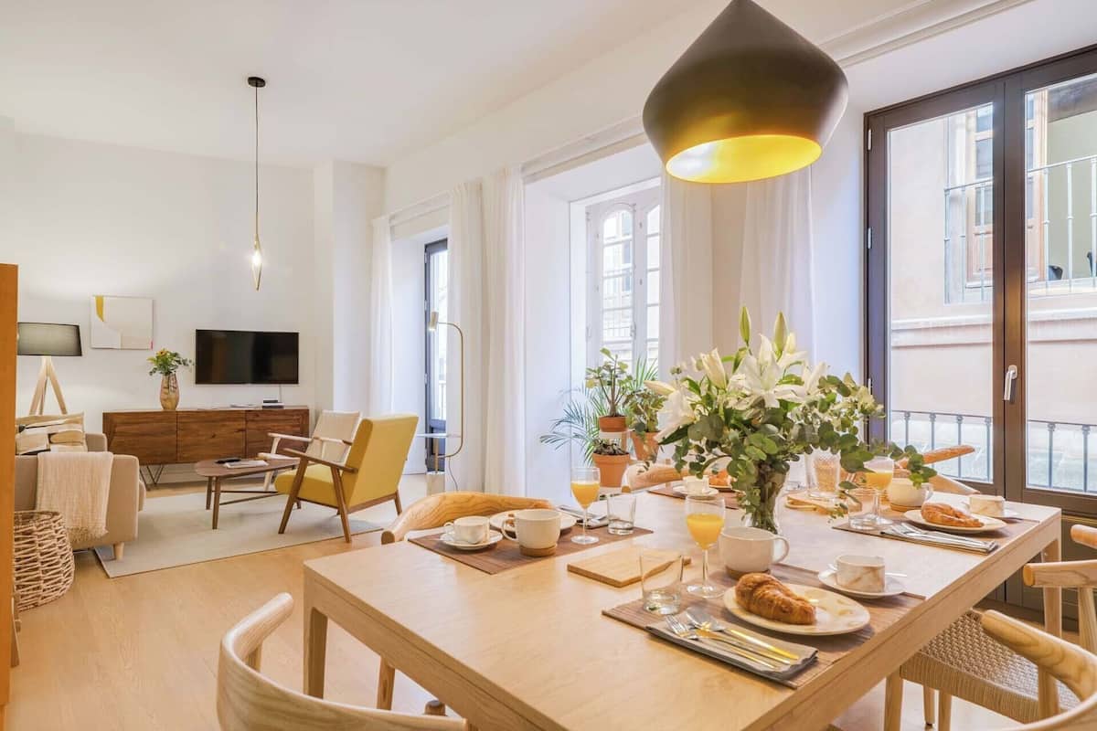 A modern dining area with a wooden table set for breakfast, a TV in the background, and a balcony with a view.