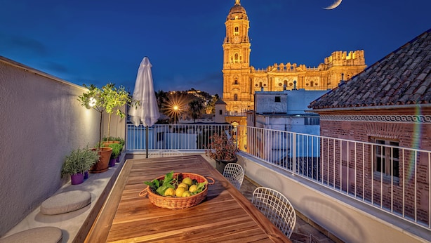 Una terrazza sul tetto con un tavolo di legno, piante in vaso e vista su un edificio storico con una torre e una luna crescente nel cielo.