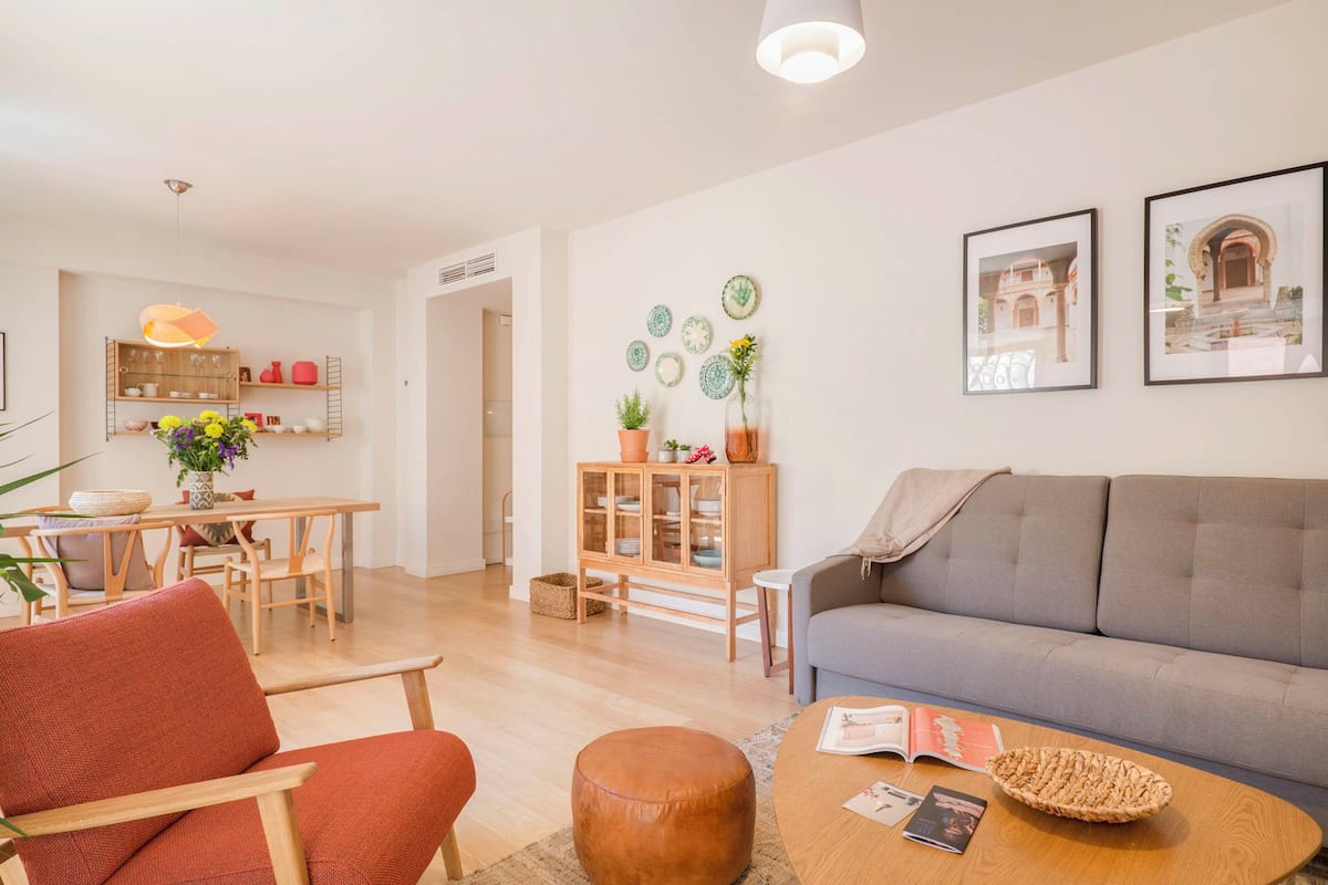 A modern living room with a grey sofa, a wooden coffee table, and a red chair. There is a dining area with a wooden table and chairs, and a kitchen area with a shelf and a hanging light.