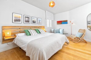 A bedroom with a wooden headboard, a large bed with white bedding, green and gray pillows, a bedside lamp, a wicker chair, a small table, and a wall-mounted shelf. - Solaga - Larios Suites (Málaga)