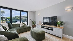 Una sala de estar moderna con un televisor grande, sofás grises, un mueble de entretenimiento de madera y vistas al exterior.