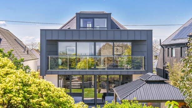 Una casa moderna de dos pisos con techo plano, ventanas grandes y un balcón visible.