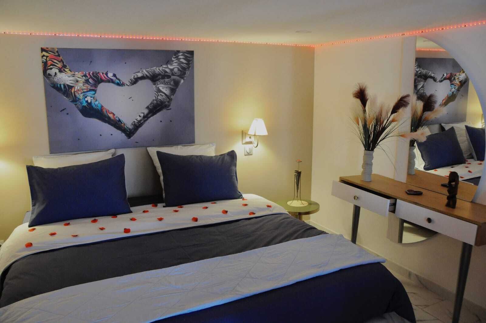 Habitación de hotel con cama, mesita de noche, espejo y un cuadro con forma de corazón en la pared.