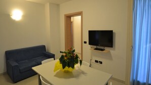 Ein Wohnzimmer mit einer Couch, einem Tisch mit einer Pflanze, einem Fernseher auf einem Schrank und einer Tür zu einem anderen Raum.