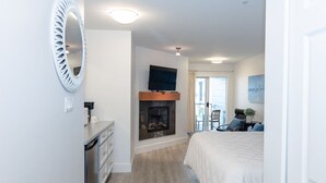A hotel room with a fireplace, a bed, a TV, and a balcony with a chair. - Ocean Breeze Seaside Studio Condo B (Nanoose Bay)