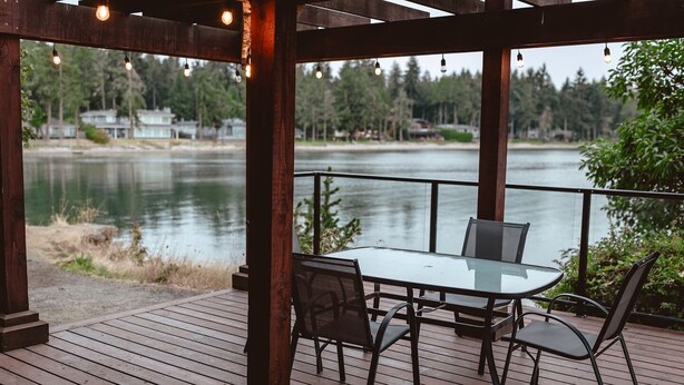 A covered outdoor deck with a table and chairs, overlooking a body of water and forested area. - Ocean Breeze Seaside Studio Condo B (Nanoose Bay)