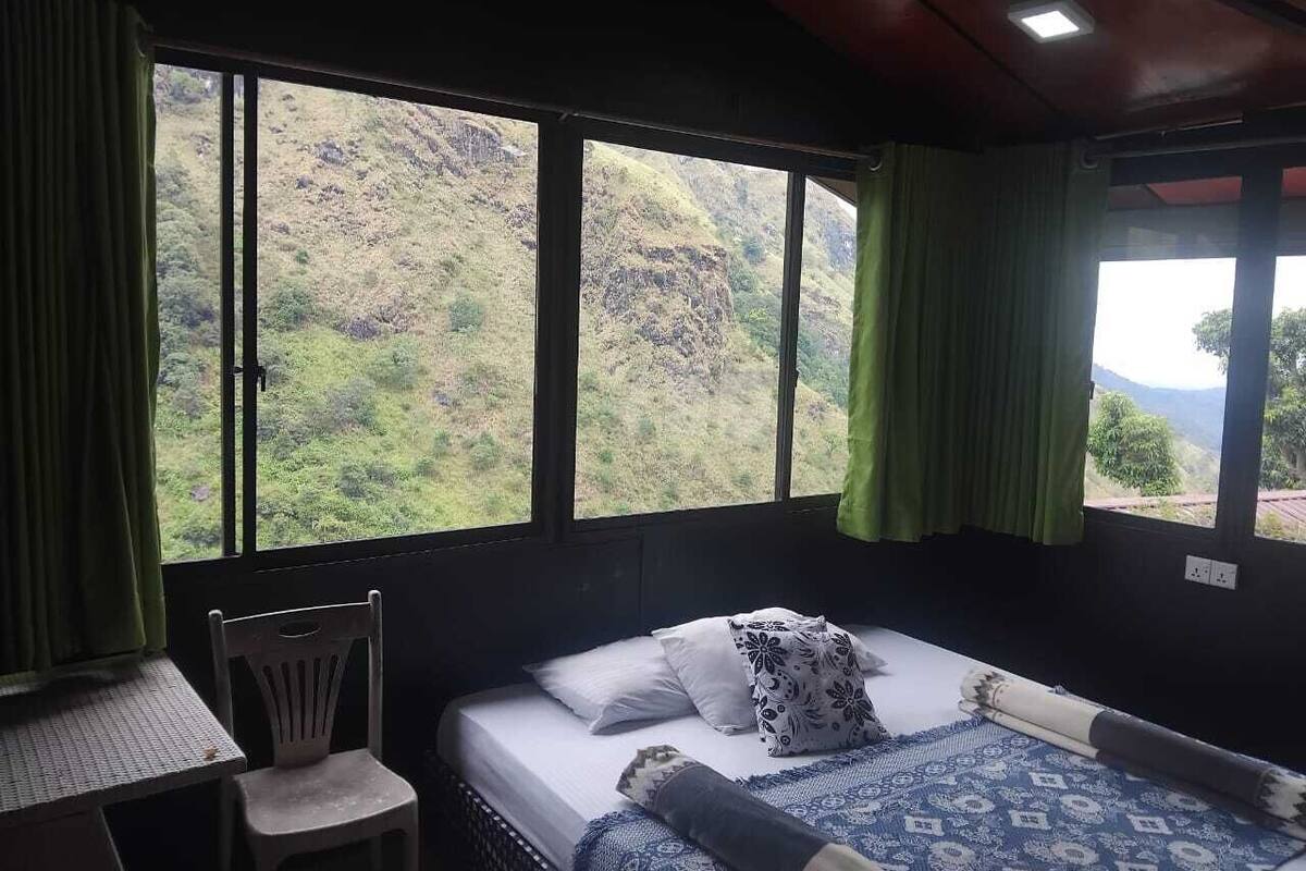 A room with a bed, a chair, and a table. There are large windows with curtains.