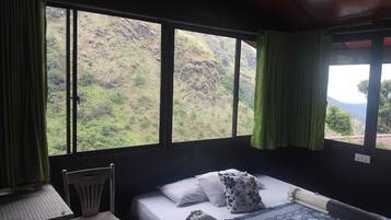 A room with a bed, a chair, and a table. There are large windows with curtains.