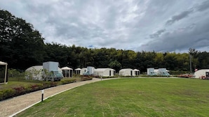 Un terrain de camping avec plusieurs camping-cars et tentes, entouré d’un champ herbeux et d’arbres.