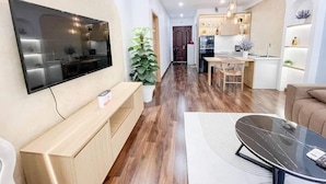 Un salon moderne doté d’un téléviseur à écran plat, d’un banc en bois, d’une table basse et d’un coin repas avec une cuisine en arrière-plan.