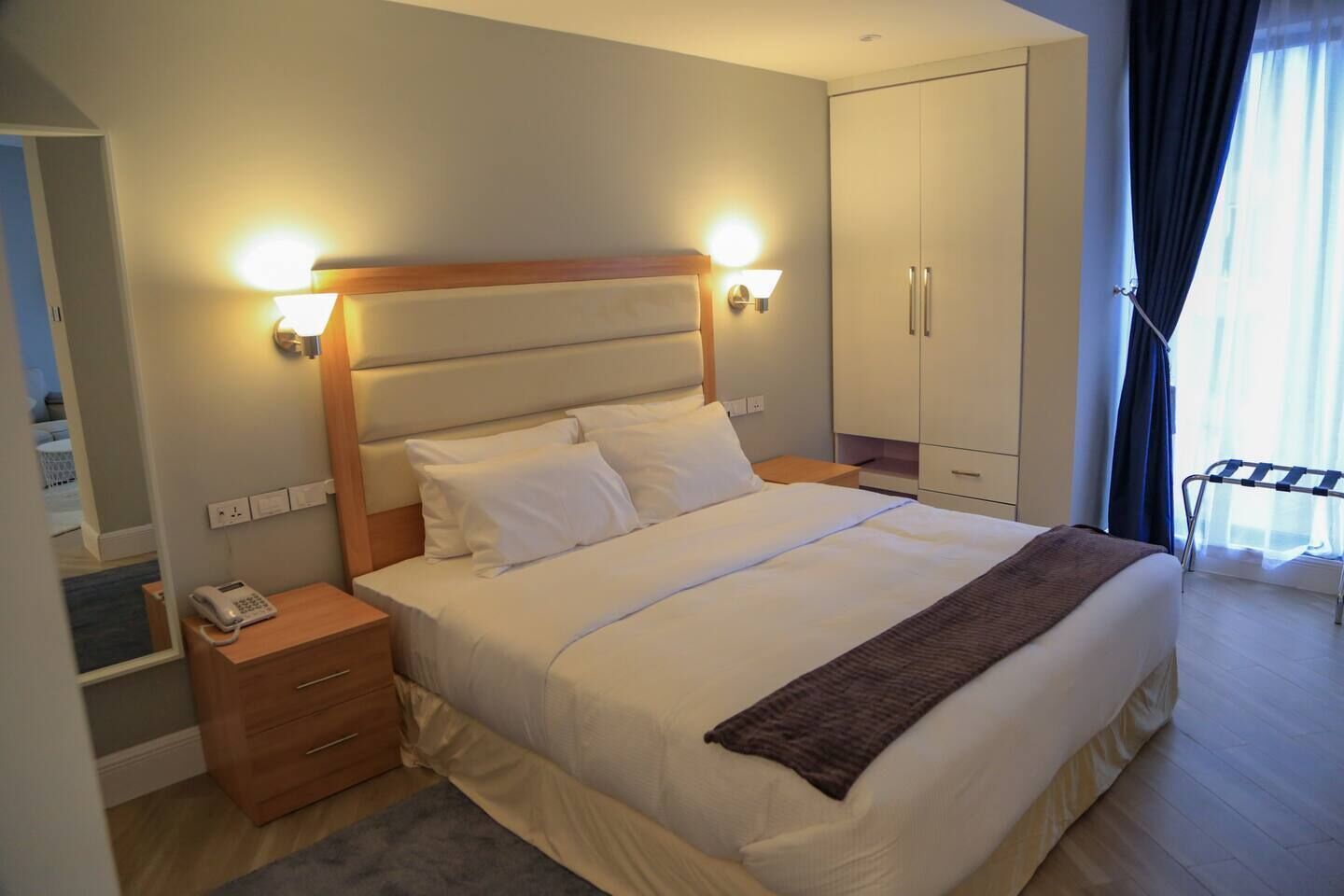 A hotel room with a large bed, bedside lamps, a wooden nightstand, a telephone, a wardrobe, and a window with curtains.