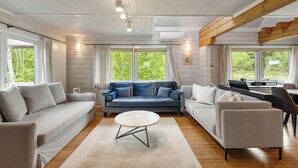 A modern living room with a blue sofa, a white sofa, and a round coffee table. There are large windows with curtains, a wooden ceiling, and an air conditioning unit. - Gekko Hanazono (Kutchan)