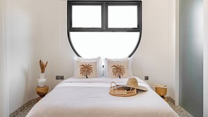 Een slaapkamer met een groot bed, twee kussens met palmboommotieven, een nachtkastje met een vaas en een mand, en een raam boven het bed.