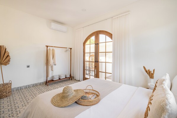 A bedroom with a large bed, a wooden wardrobe, a window with curtains, and a small table with a basket and a hat. - LA LUNA (Canggu)