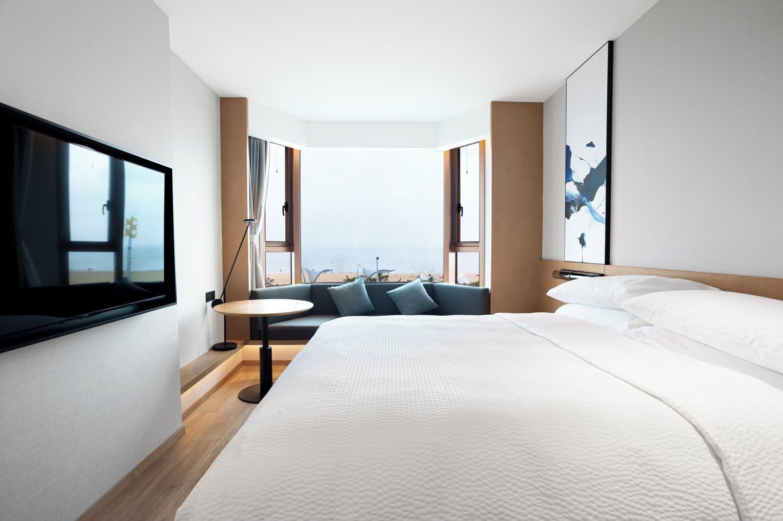 Ett modernt hotellrum med en stor säng, en tv som är monterad på väggen, ett litet runt bord och en soffa med utsikt över stranden.