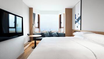 Ett modernt hotellrum med en stor säng, en tv som är monterad på väggen, ett litet runt bord och en soffa med utsikt över stranden.