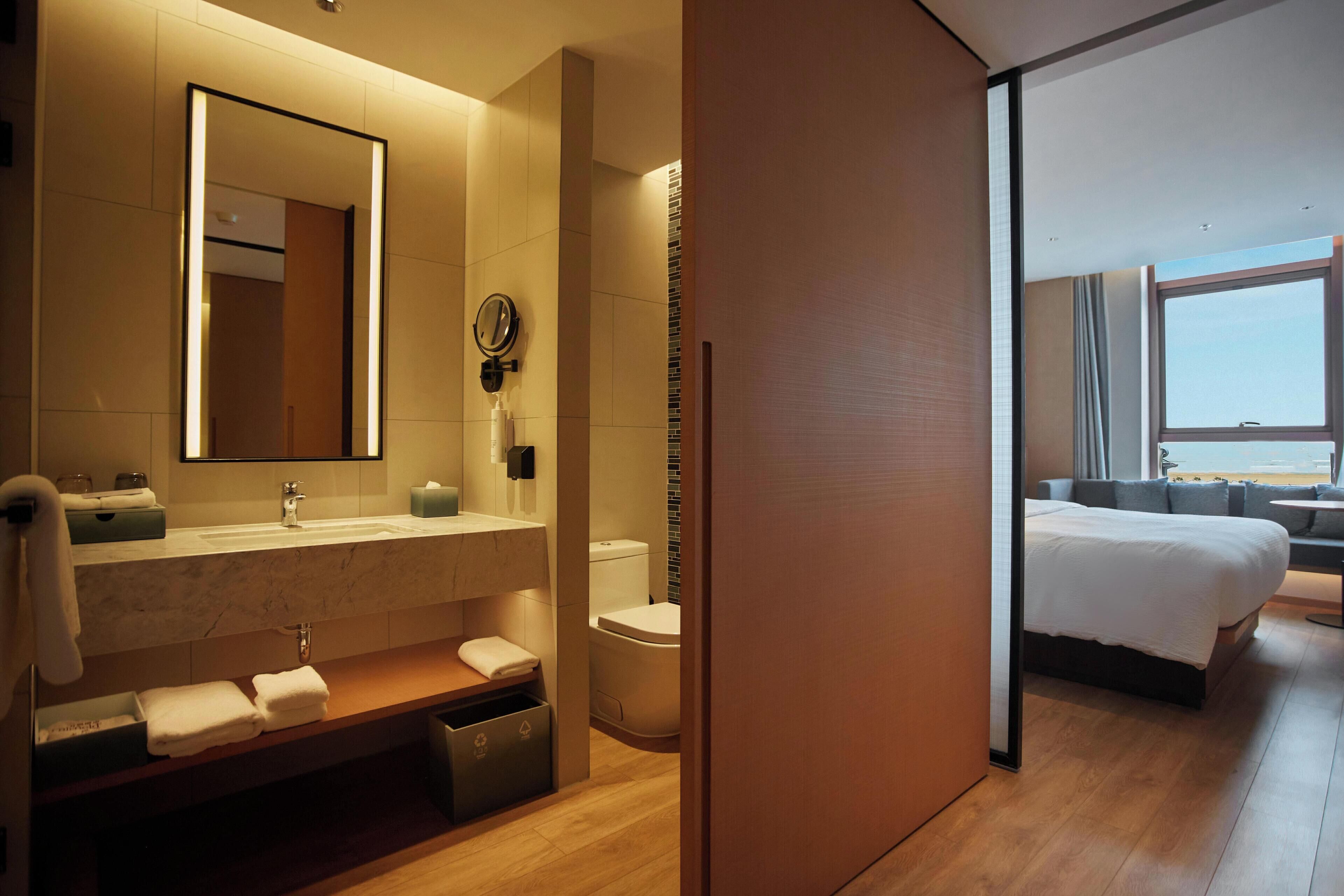 Un moderno baño de hotel con un espejo grande, lavabo y retrete. Hay un banco de madera con toallas y un cubo de basura. El cuarto tiene piso de madera y un ventanal con vistas.