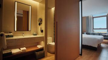 A modern hotel bathroom with a large mirror, sink, and toilet. There is a wooden bench with towels and a trash bin. The room has a wooden floor and a large window with a view.