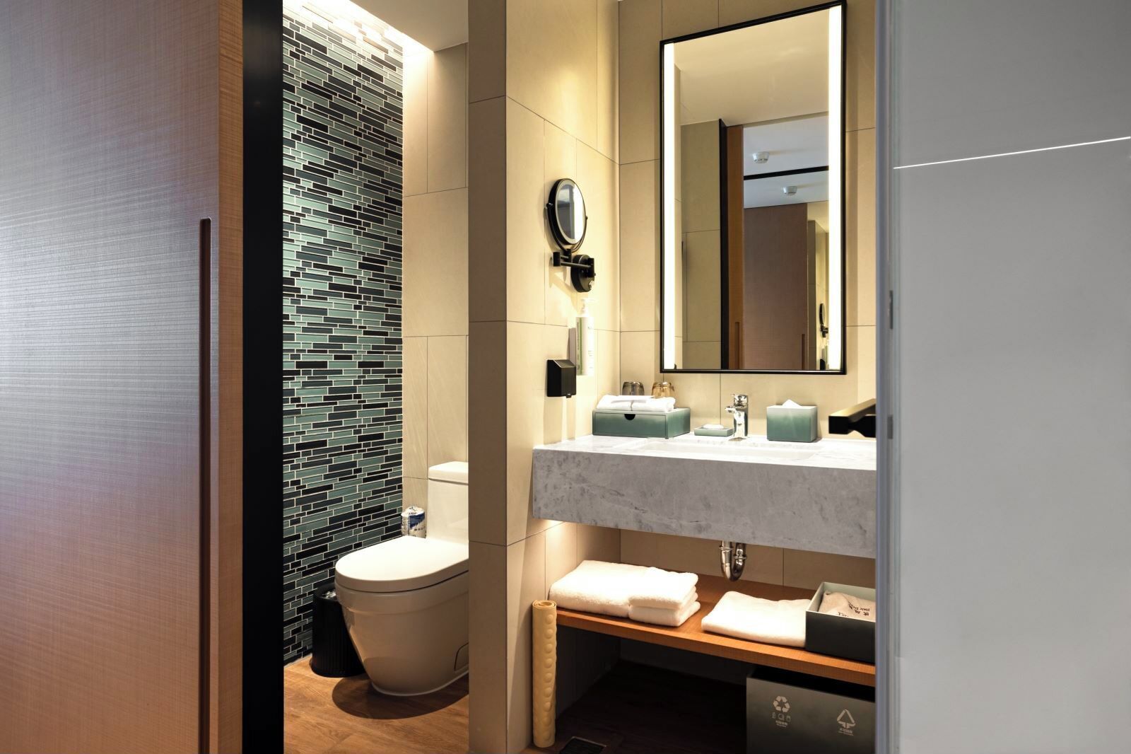 A modern bathroom with a tiled wall, a toilet, a sink with a mirror, and a wooden bench.