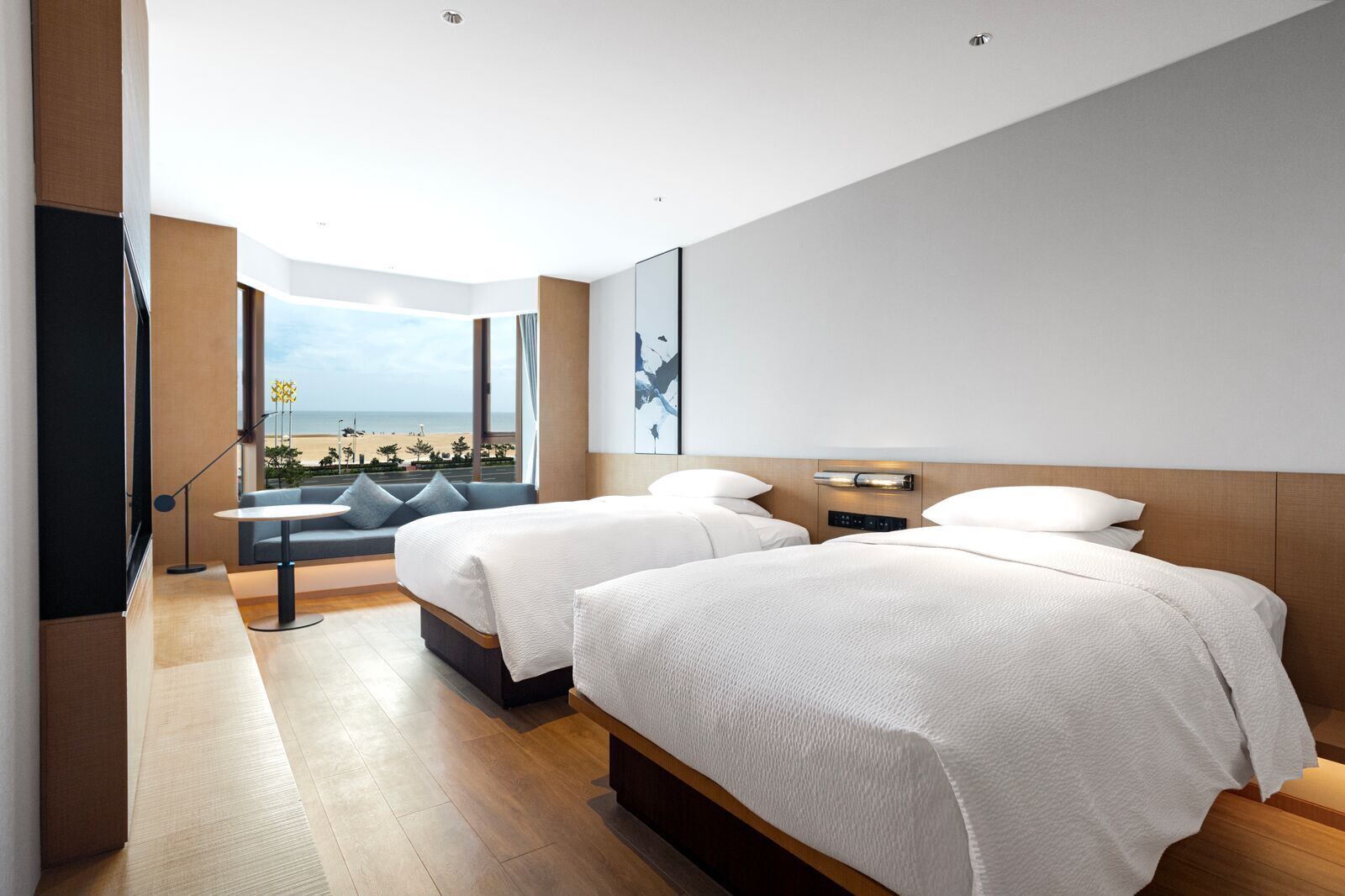 A modern hotel room with two beds, a large window with a beach view, and a seating area with a view.