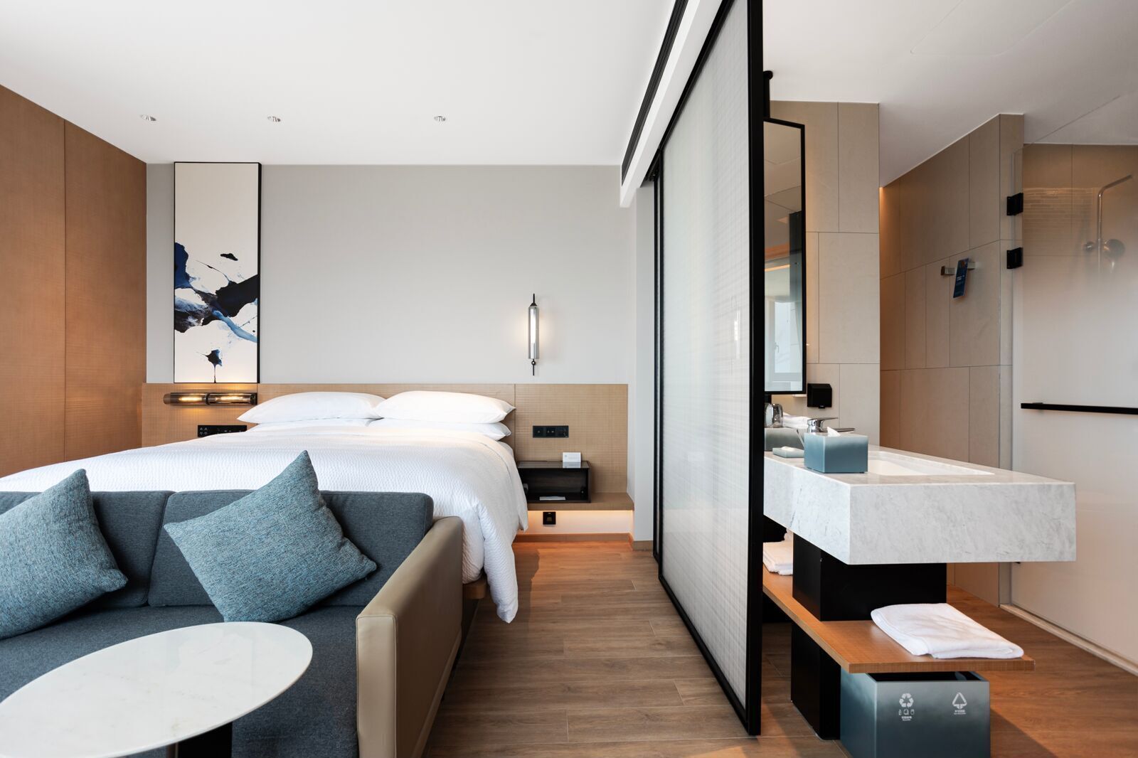 A modern hotel room with a bed, a sofa, a small table, a bathroom with a sink and mirror, and a shower area.