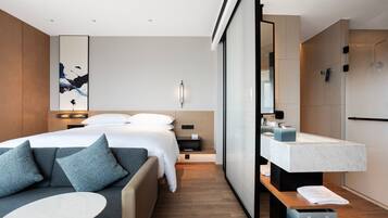 A modern hotel room with a bed, a sofa, a small table, a bathroom with a sink and mirror, and a shower area.