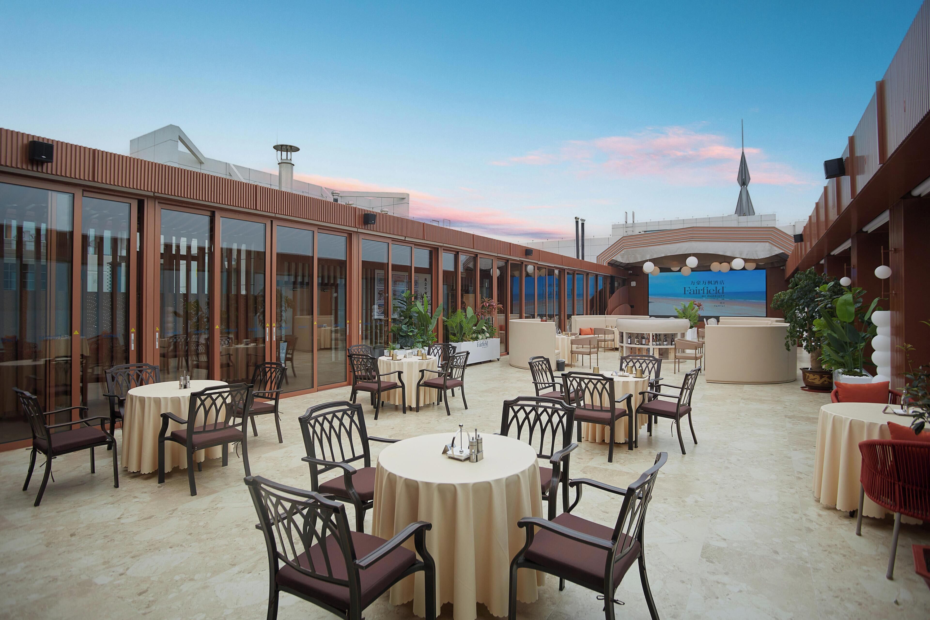 An outdoor dining area with round tables and chairs, surrounded by glass walls and potted plants.