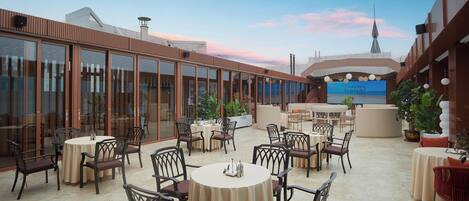 An outdoor dining area with round tables and chairs, surrounded by glass walls and potted plants.